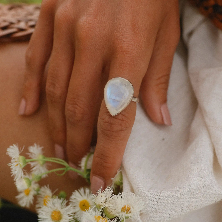 Woman's hand holding daisies wearing a teardrop shaped rainbow moonstone ring.