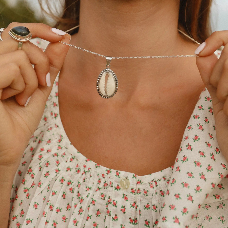woman wearing white top with red flowers holding up a sterling silver necklace with a cowrie sea shell - shop women’s cowrie sea shell jewellery Australia 