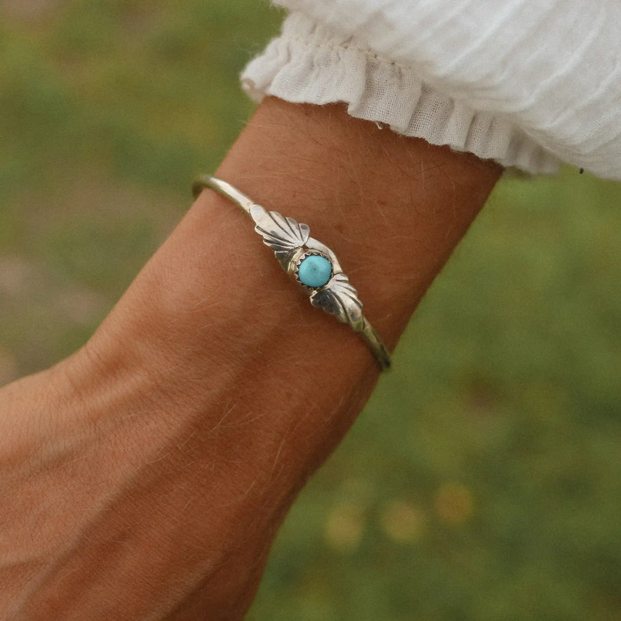 Woman wearing a white top and a silver cuff with a small turquoise stone on it and feather-like detailing engraved in the silver on either side.