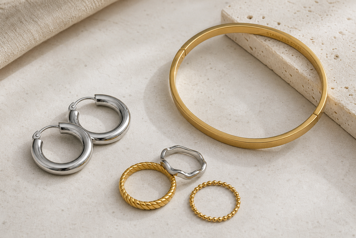 Stainless steel hoop earrings, a gold bangle, and stacker rings arranged on a neutral surface