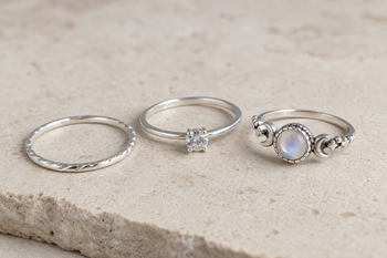 Three women's promise rings on a neutral surface: a slim silver band, a gemstone solitaire ring, and a moonstone ring.