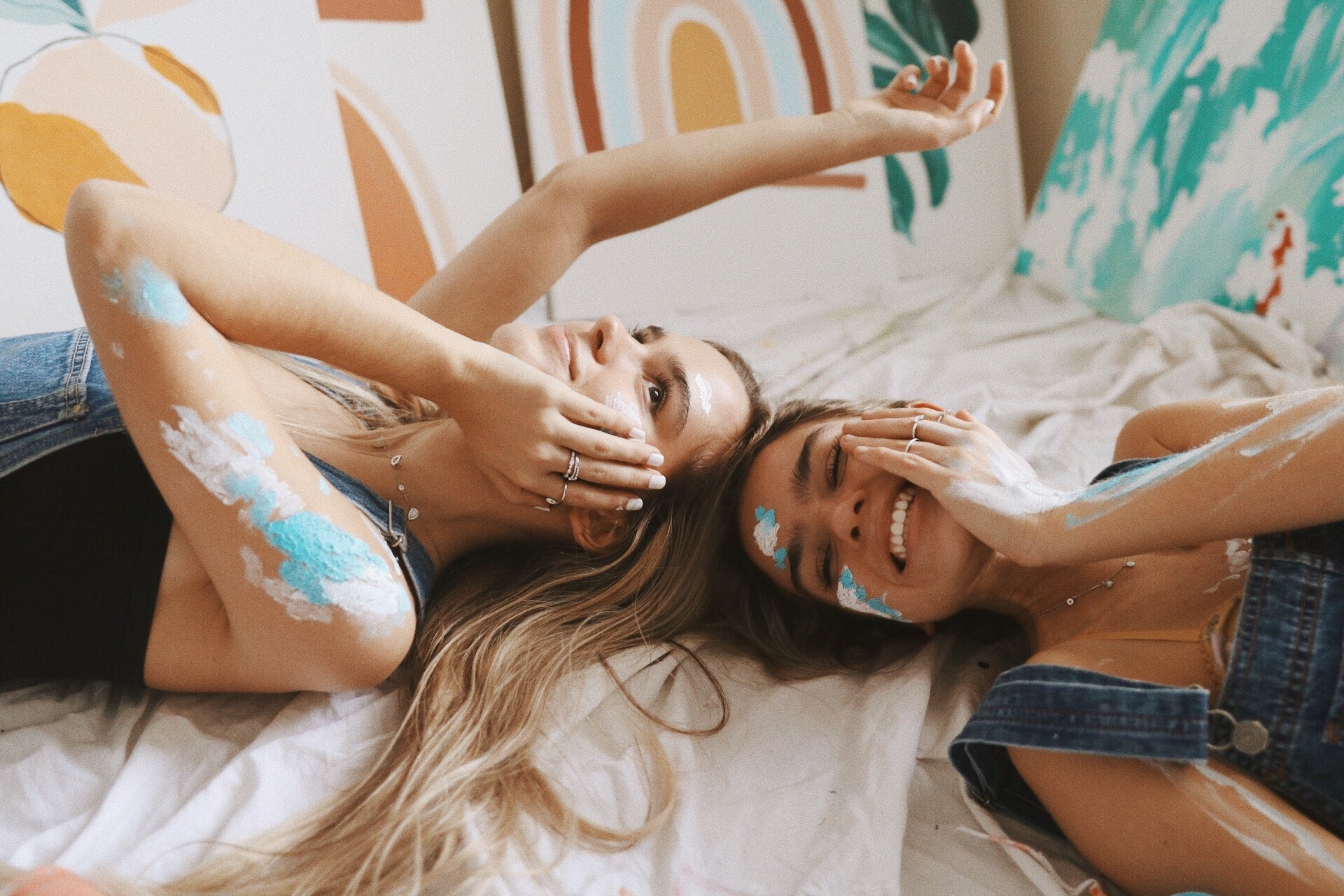 Two women lying on a bed with paint on their arms, surrounded by colorful pillows.