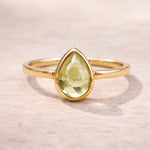 Gold ring with a teardrop-shaped green natural peridot gemstone on a light wooden background
