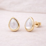 Gold stud earrings with teardrop-shaped white pearls on a light pink background