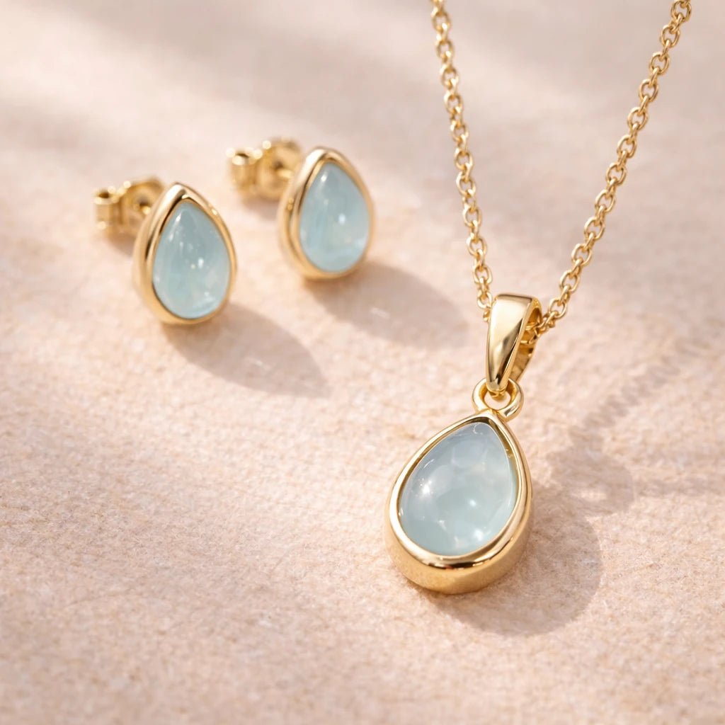 Gold necklace and stud earrings with light blue natural aquamarine teardrop stones on a soft pink background