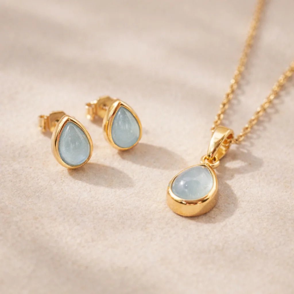 Gold Jewellery set with pear drop-shaped pendant and earrings with natural bezel set aquamarine gemstones on a soft pink background