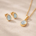 Gold Jewellery set with pear drop-shaped pendant and earrings with natural bezel set aquamarine gemstones on a soft pink background