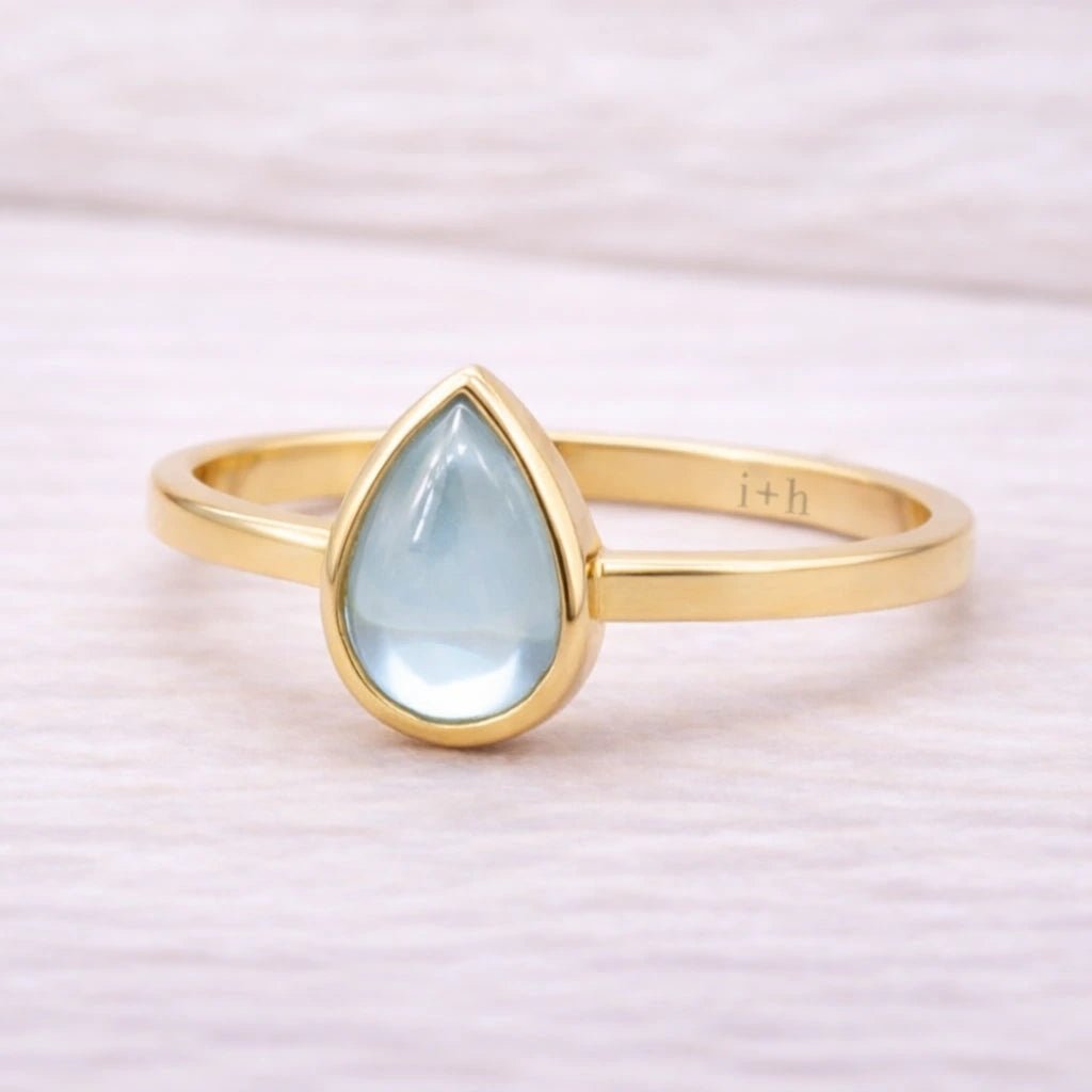 Gold ring with a teardrop-shaped light blue natural topaz gemstone on a soft pink background