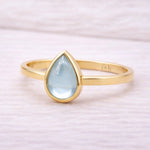 Gold ring with a teardrop-shaped light blue natural topaz gemstone on a soft pink background