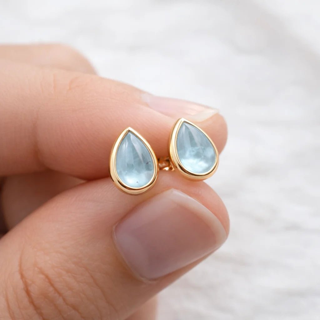Gold teardrop earrings with light blue natural aquamarine gemstones held between fingers against a neutral background