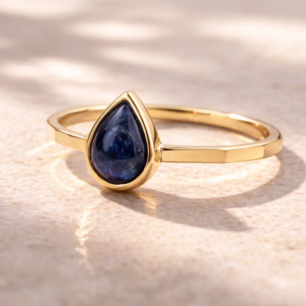 Gold ring with a blue pear shaped natural sapphire gemstone on a textured beige background