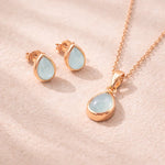 Rose Gold Jewellery set with pear drop-shaped pendant and earrings with natural bezel set aquamarine gemstones on a soft pink background