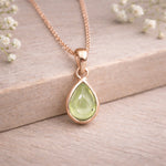 Rose gold necklace with a green teardrop pendant on a wooden surface with white flowers.