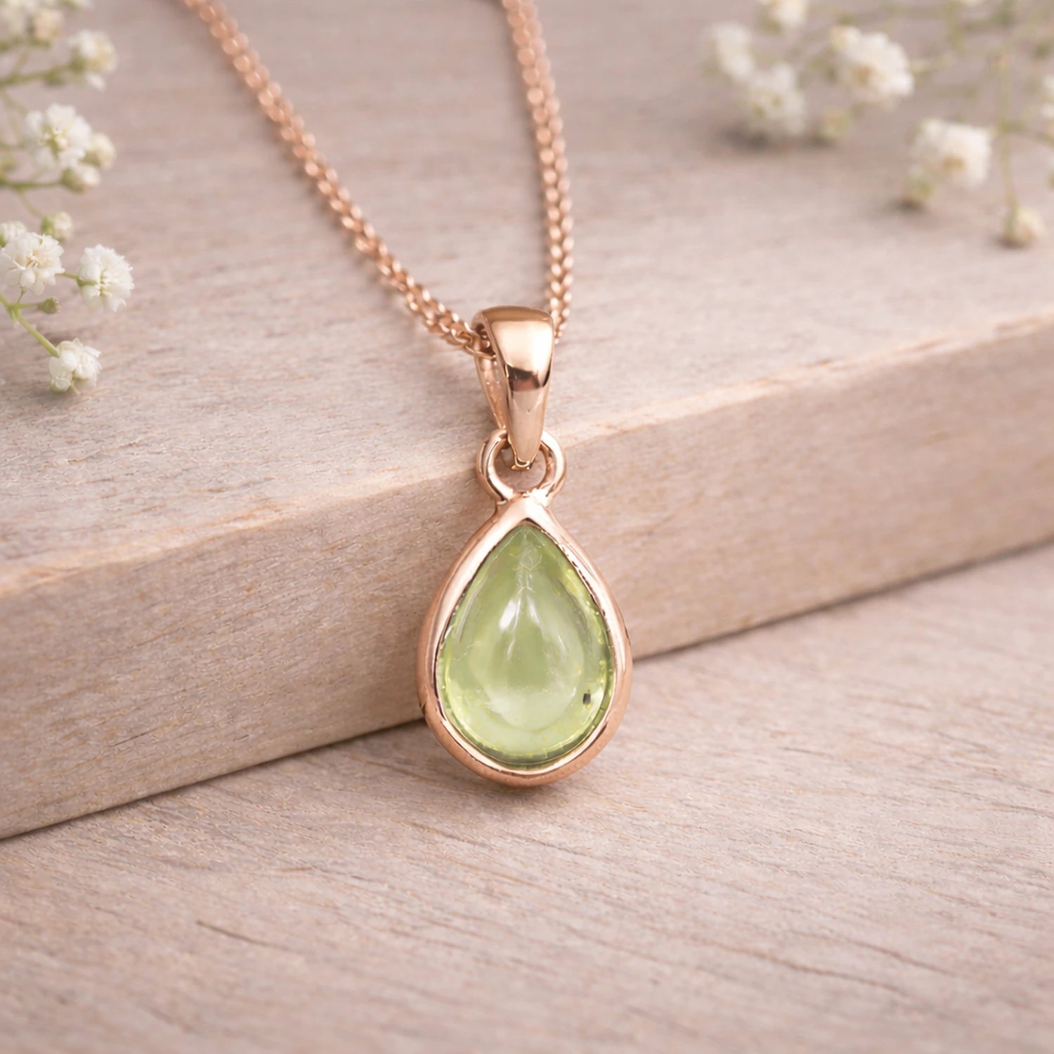Rose gold necklace with a green teardrop pendant on a wooden surface with white flowers.