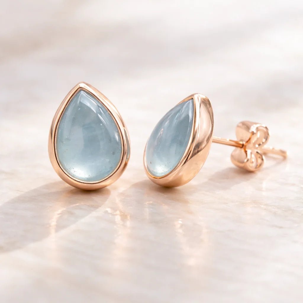 Rose gold earrings with teardrop-shaped light blue natural aquamarine gemstones on a soft beige background