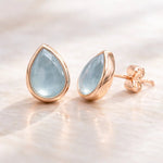 Rose gold earrings with teardrop-shaped light blue natural aquamarine gemstones on a soft beige background