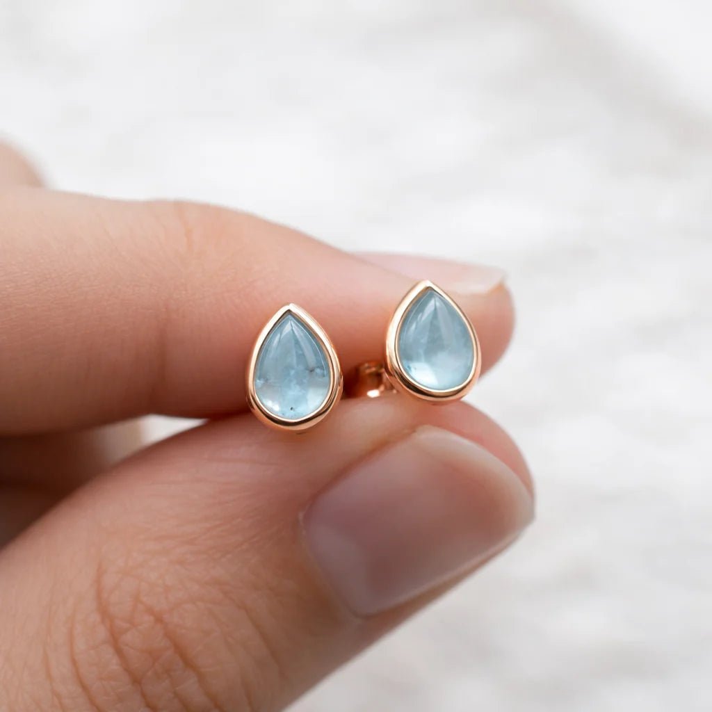 Teardrop-shaped aquamarine gemstone earrings with rose gold setting held between fingers on a white background