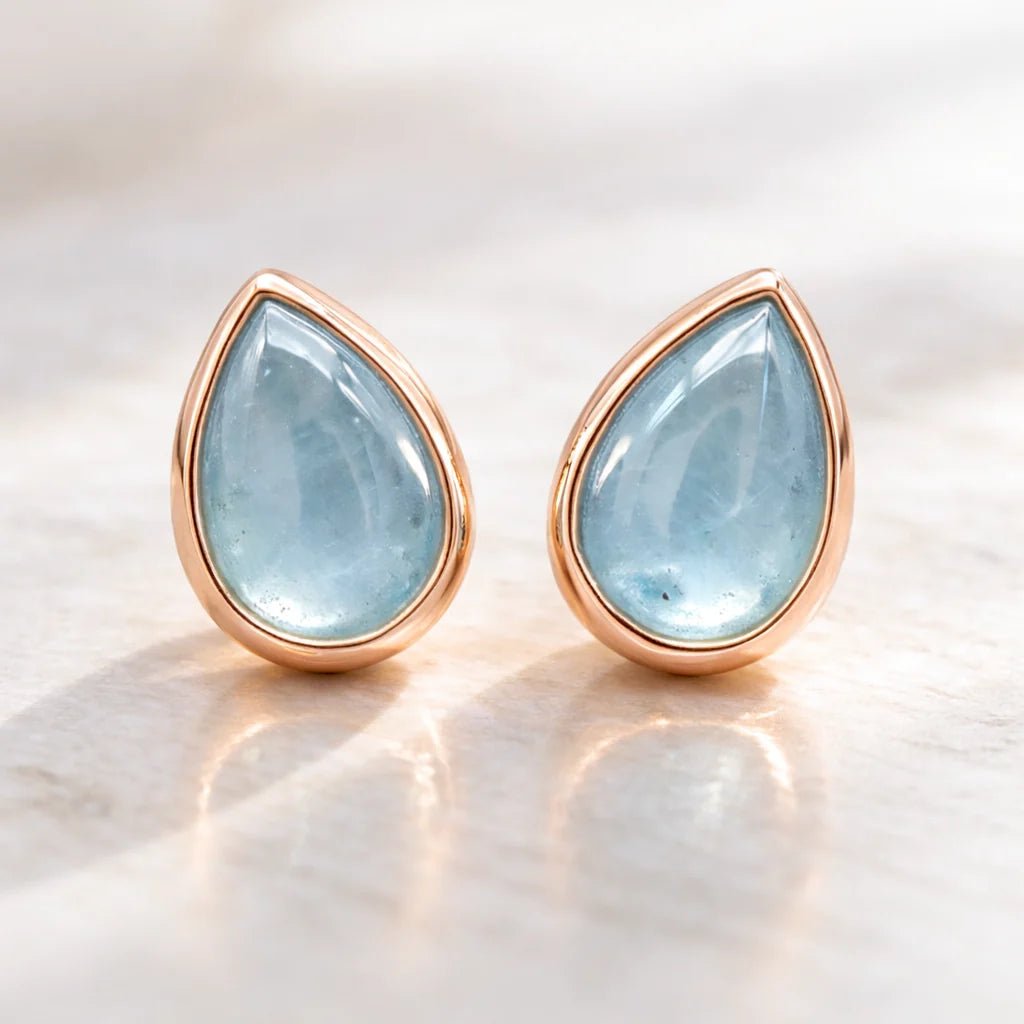 Teardrop-shaped earrings with rose gold frames and light blue aquamarine gemstones on a marble surface.