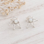 A pair of silver butterfly stud earrings with oval white opal centers, displayed on a light, rustic wooden surface with dried flowers in the background.
