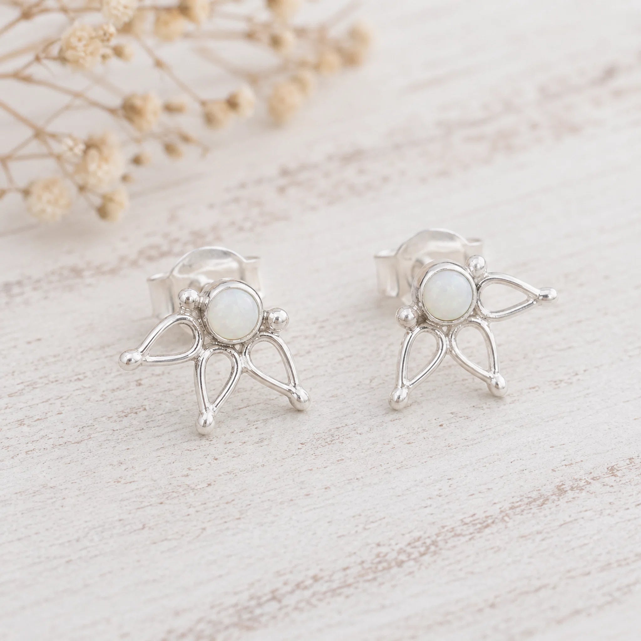 A pair of silver butterfly stud earrings with oval white opal centers, displayed on a light, rustic wooden surface with dried flowers in the background.