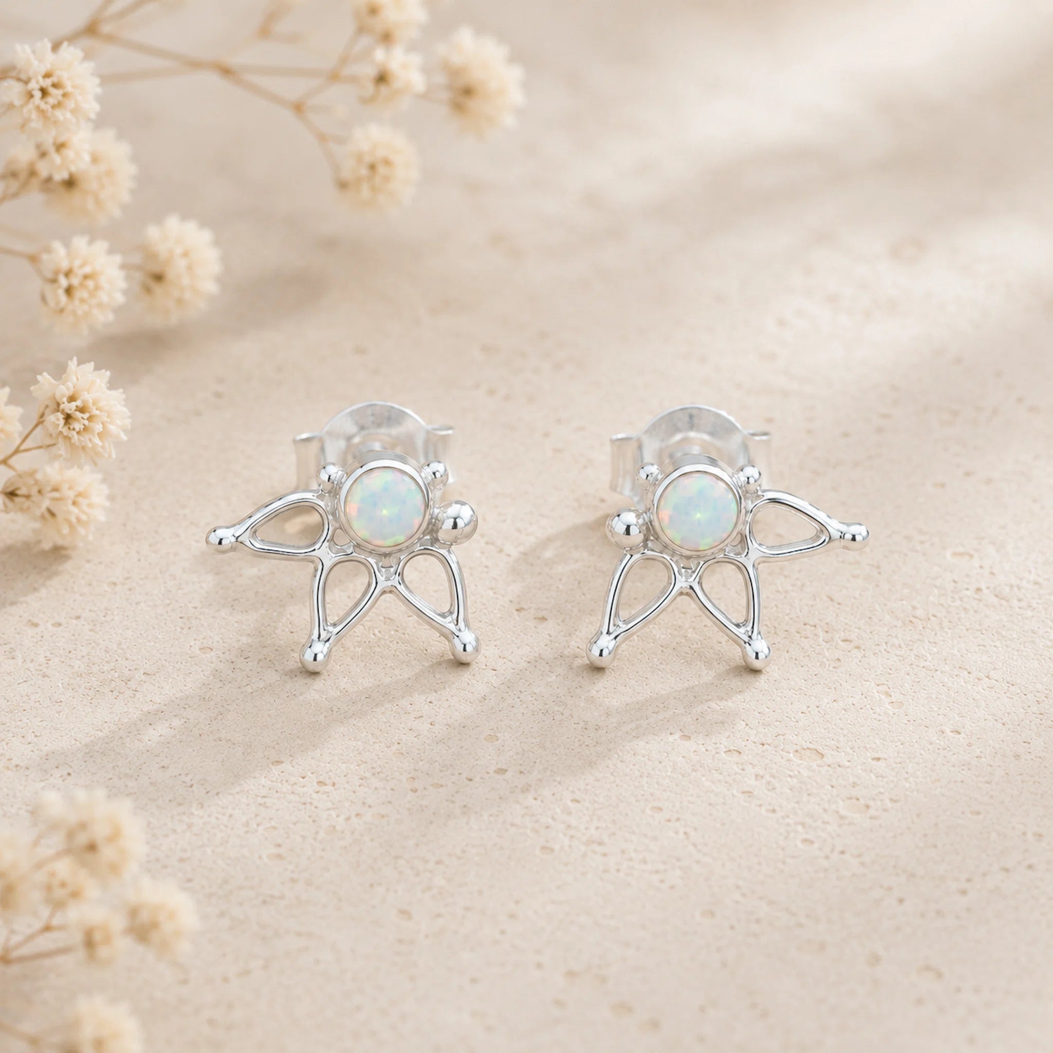 A pair of silver stud earrings with a delicate openwork starburst design surrounding a round iridescent white opal center, displayed on a textured beige surface with dried flowers.