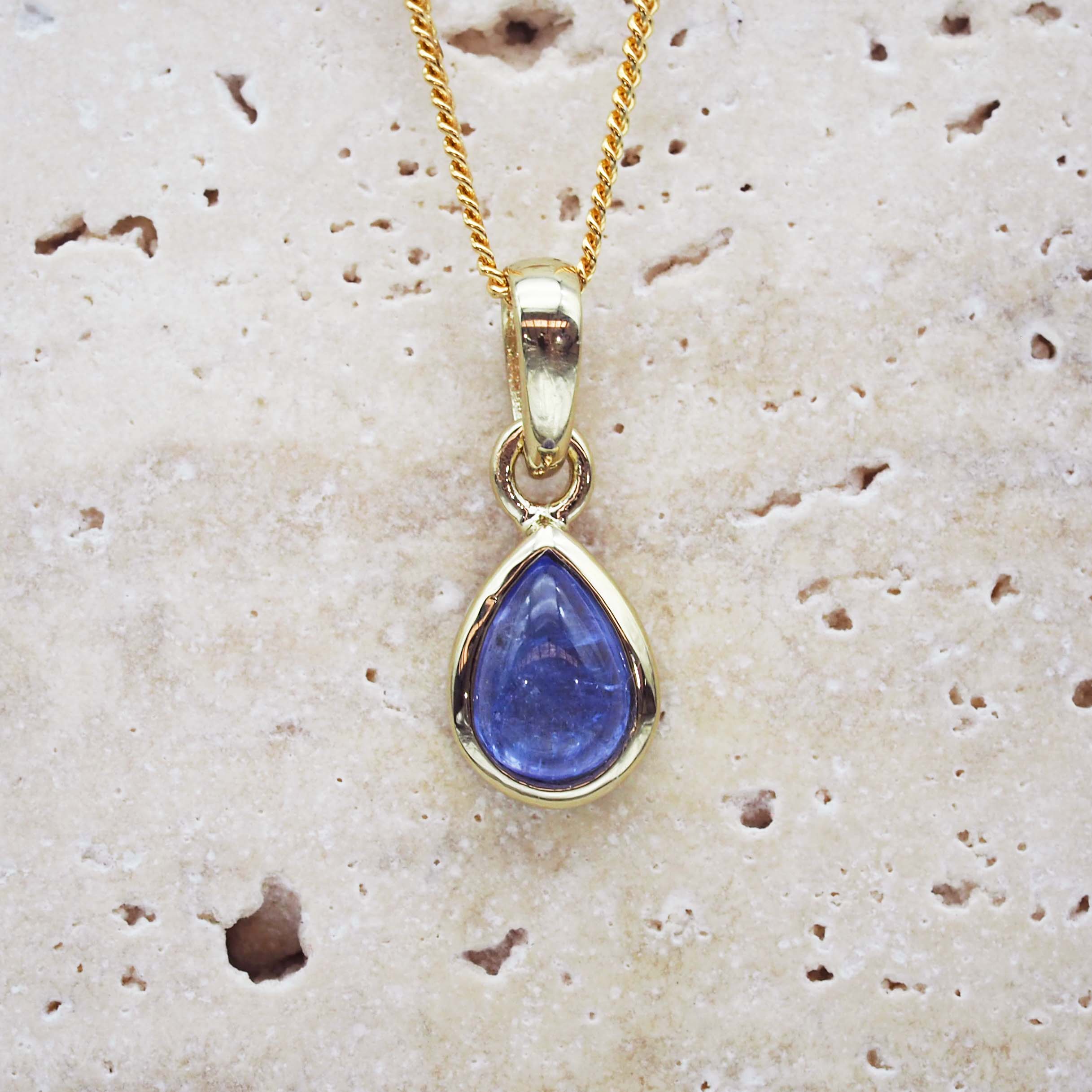 Gold pendant with a blue teardrop-shaped gemstone on a textured beige surface