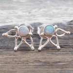A pair of silver stud earrings featuring a small iridescent opal gemstone in a circular setting, with three delicate branching silver prongs below, displayed on a rustic wooden surface.