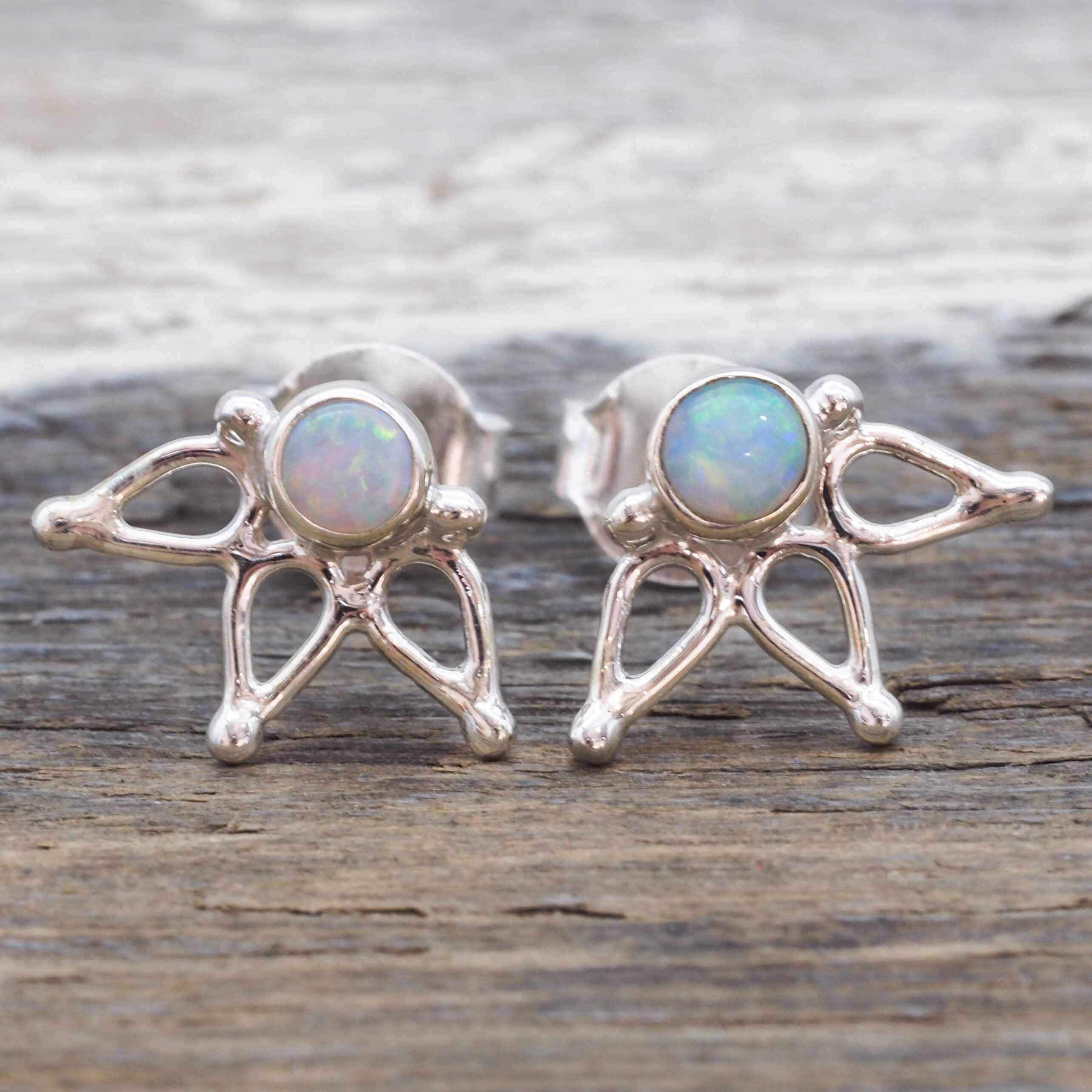 A pair of silver stud earrings featuring a small iridescent opal gemstone in a circular setting, with three delicate branching silver prongs below, displayed on a rustic wooden surface.
