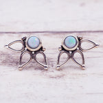 A pair of silver-toned winged fairy stud earrings with round iridescent opal gemstones, displayed on a light whitewashed wooden surface.
