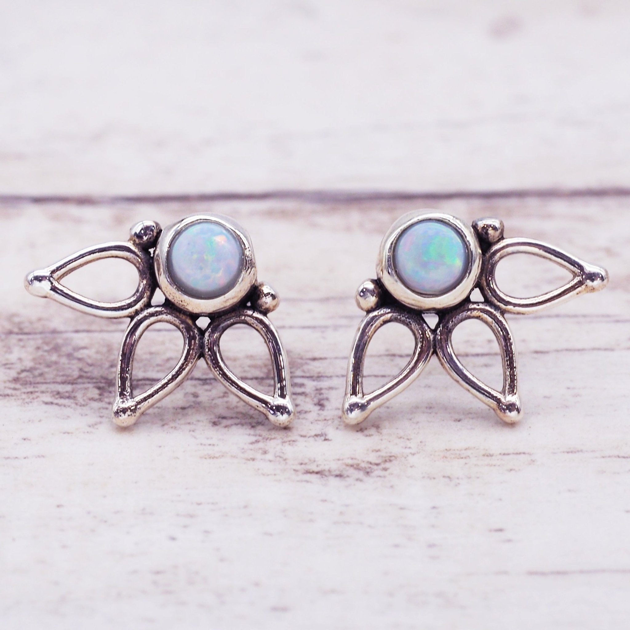 A pair of silver-toned winged fairy stud earrings with round iridescent opal gemstones, displayed on a light whitewashed wooden surface.