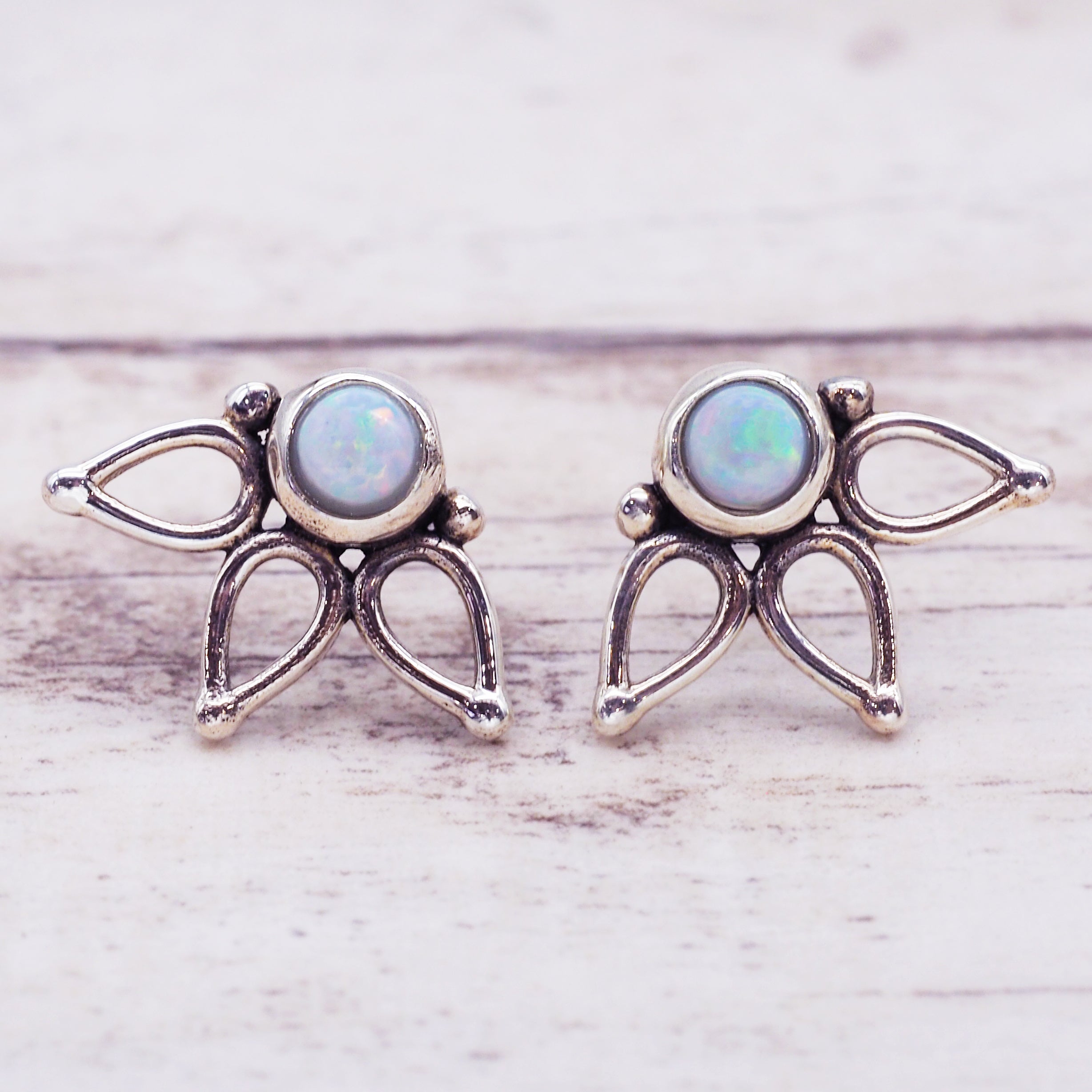 A pair of silver-toned winged fairy stud earrings with round iridescent opal gemstones, displayed on a light whitewashed wooden surface.