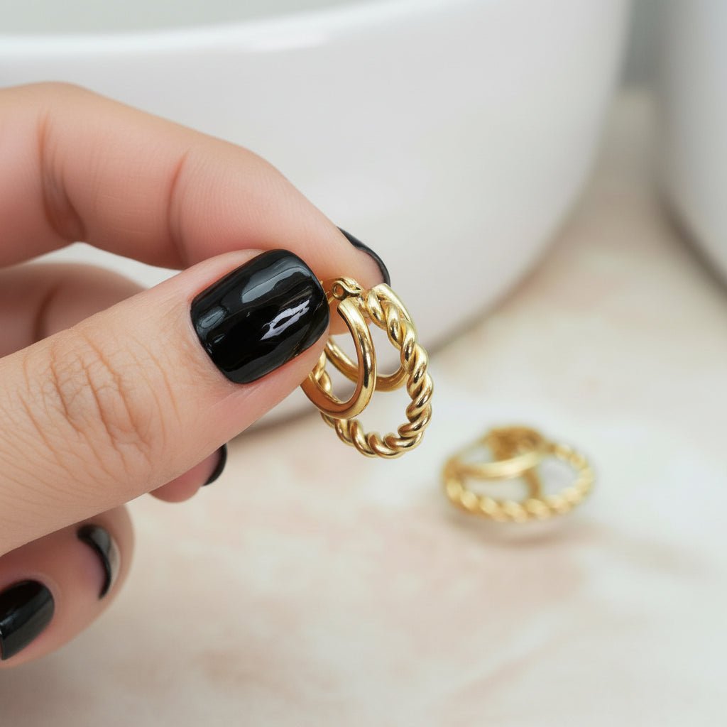 Gold earrings being held by a hand with black nail polish on a neutral background