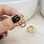 Gold earrings being held by a hand with black nail polish on a neutral background
