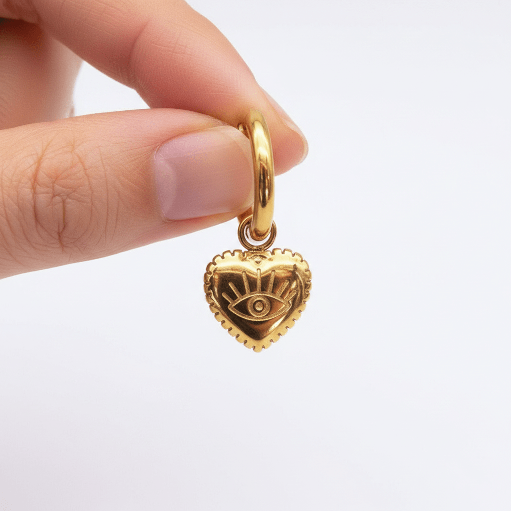 Gold hoop earring with a heart-shaped charm featuring an eye design, held by a hand against a white background.