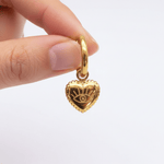 Gold hoop earring with a heart-shaped charm featuring an eye design, held by a hand against a white background.