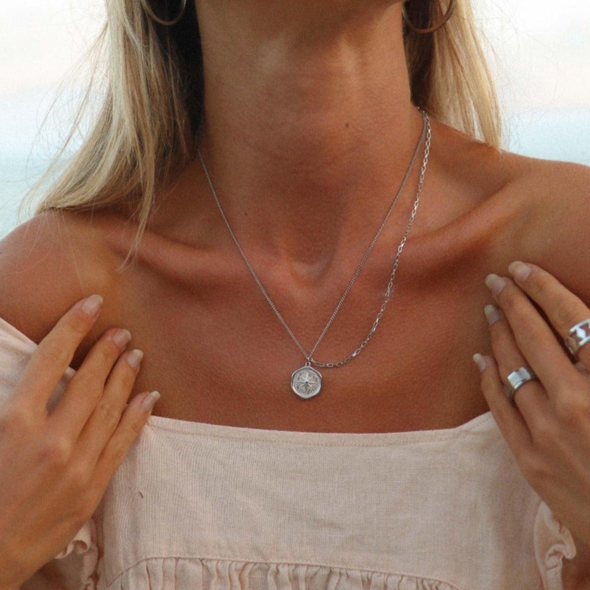 Close-up of a woman wearing a silver necklace with a coin pendant, with a blurred background.