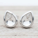 A pair of silver teardrop stud earrings with clear quartz faceted gemstones, resting on a light-colored wooden surface.