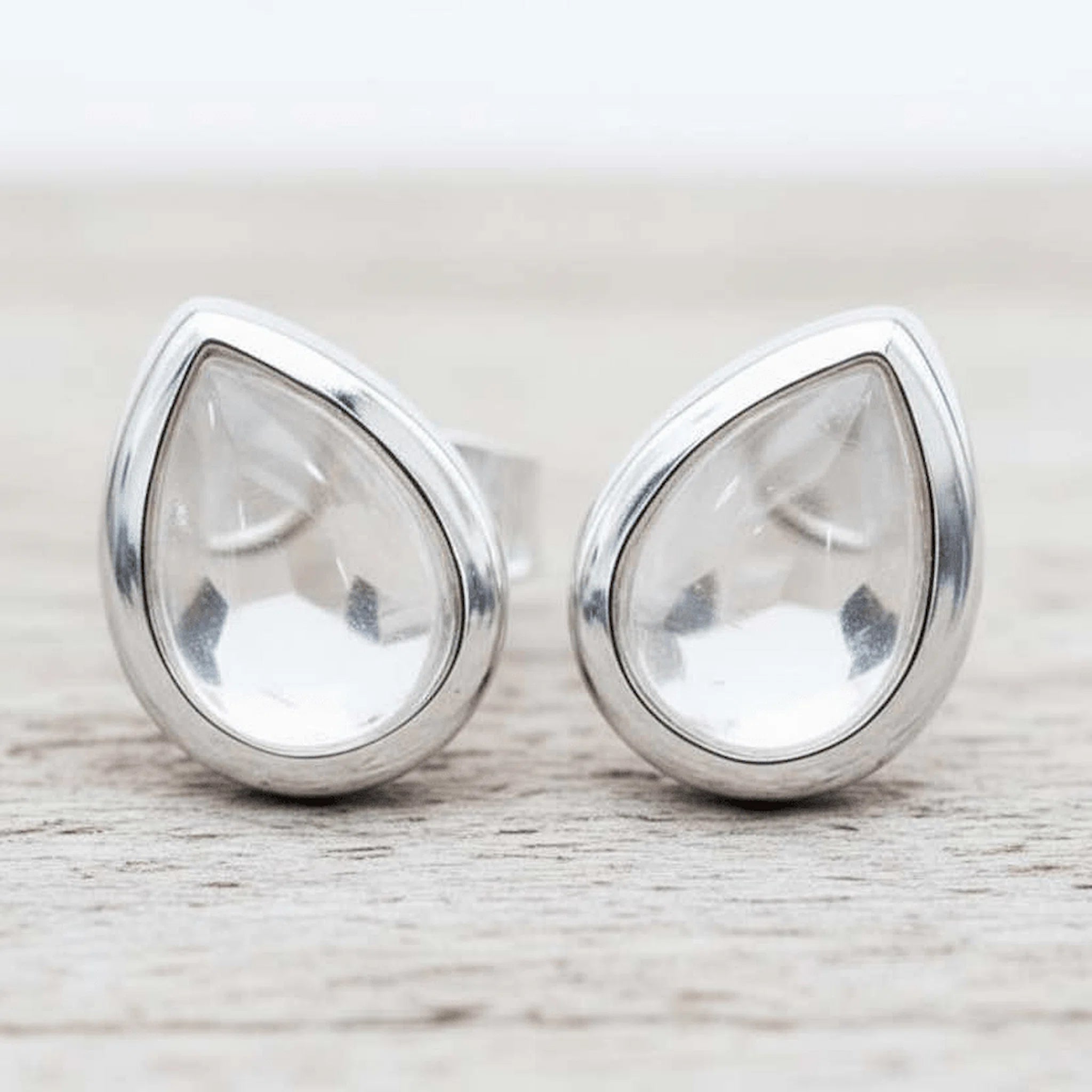 A pair of silver teardrop stud earrings with clear quartz faceted gemstones, resting on a light-colored wooden surface.