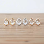 Six teardrop-shaped clear gemstone stud earrings in gold, silver, and rose gold settings, lined up on a light wooden surface.