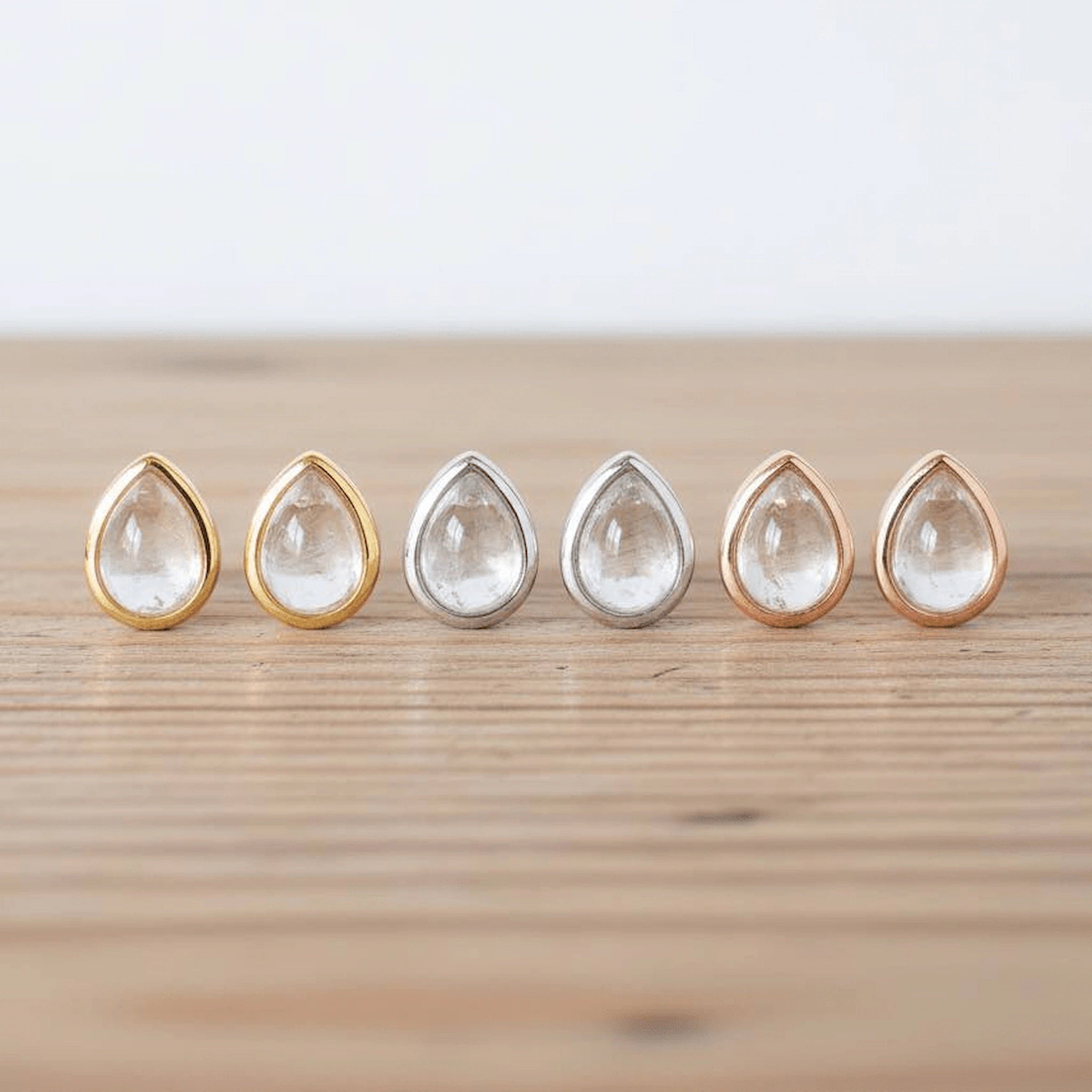 Six teardrop-shaped clear gemstone stud earrings in gold, silver, and rose gold settings, lined up on a light wooden surface.