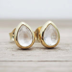Teardrop-shaped clear quartz gemstone stud earrings in gold settings, resting on a light gray wooden surface.