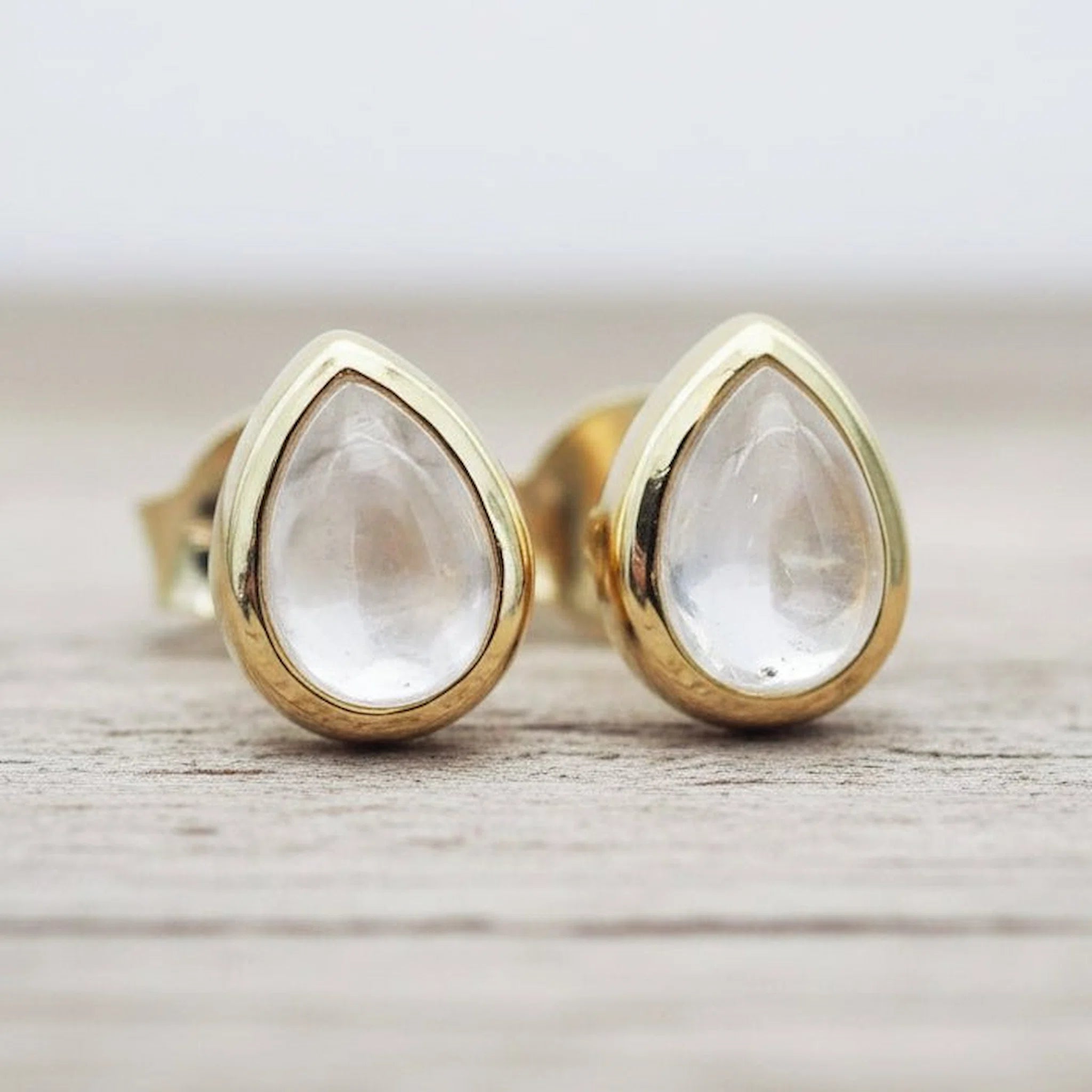 Teardrop-shaped clear quartz gemstone stud earrings in gold settings, resting on a light gray wooden surface.