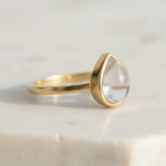 Gold ring with a clear herkimer quartz teardrop-shaped gemstone on a light background