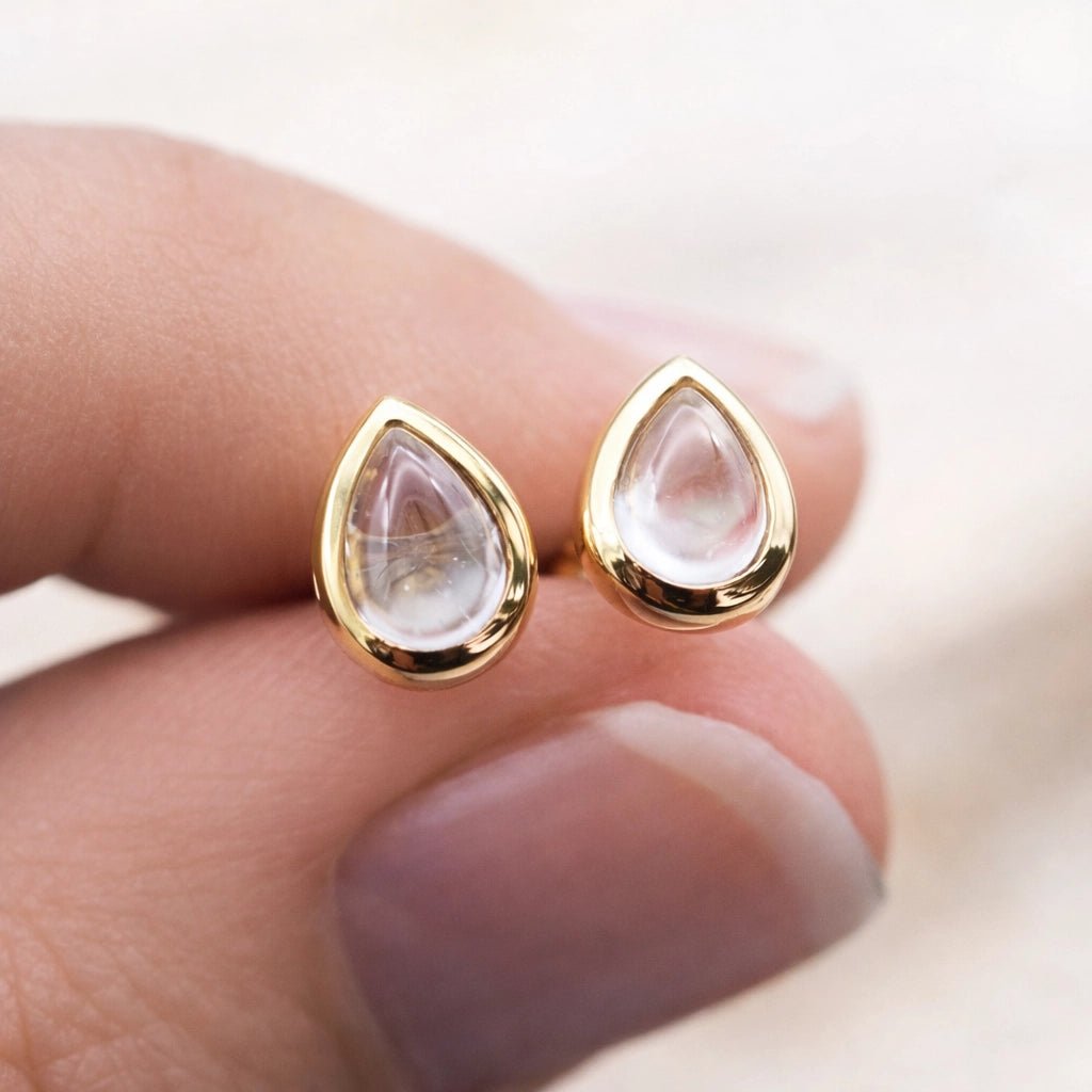 Gold teardrop stud earrings with clear natural herkimer quartz stones on a hand against a blurred background