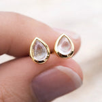 Gold teardrop stud earrings with clear natural herkimer quartz stones on a hand against a blurred background