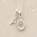 Sterling Silver double pendant necklace with a teardrop-shaped april birthstone clear quartz pendant and letter 'A' charm on a beige background