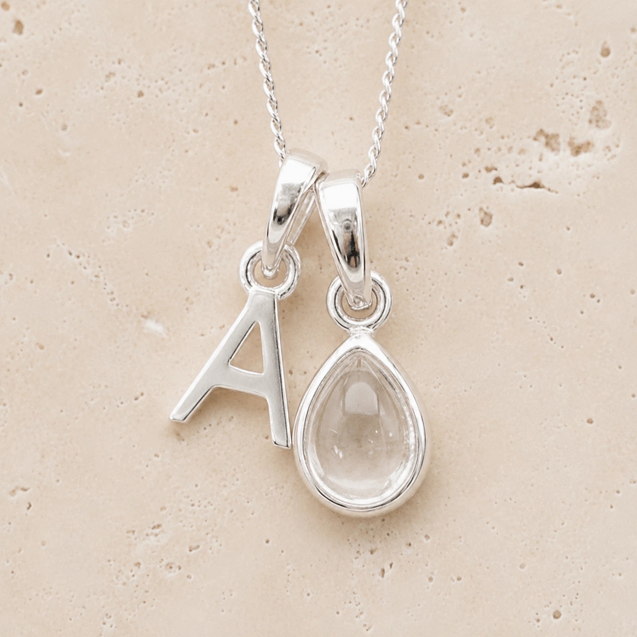 Sterling Silver double pendant necklace with a teardrop-shaped april birthstone clear quartz pendant and letter 'A' charm on a beige background