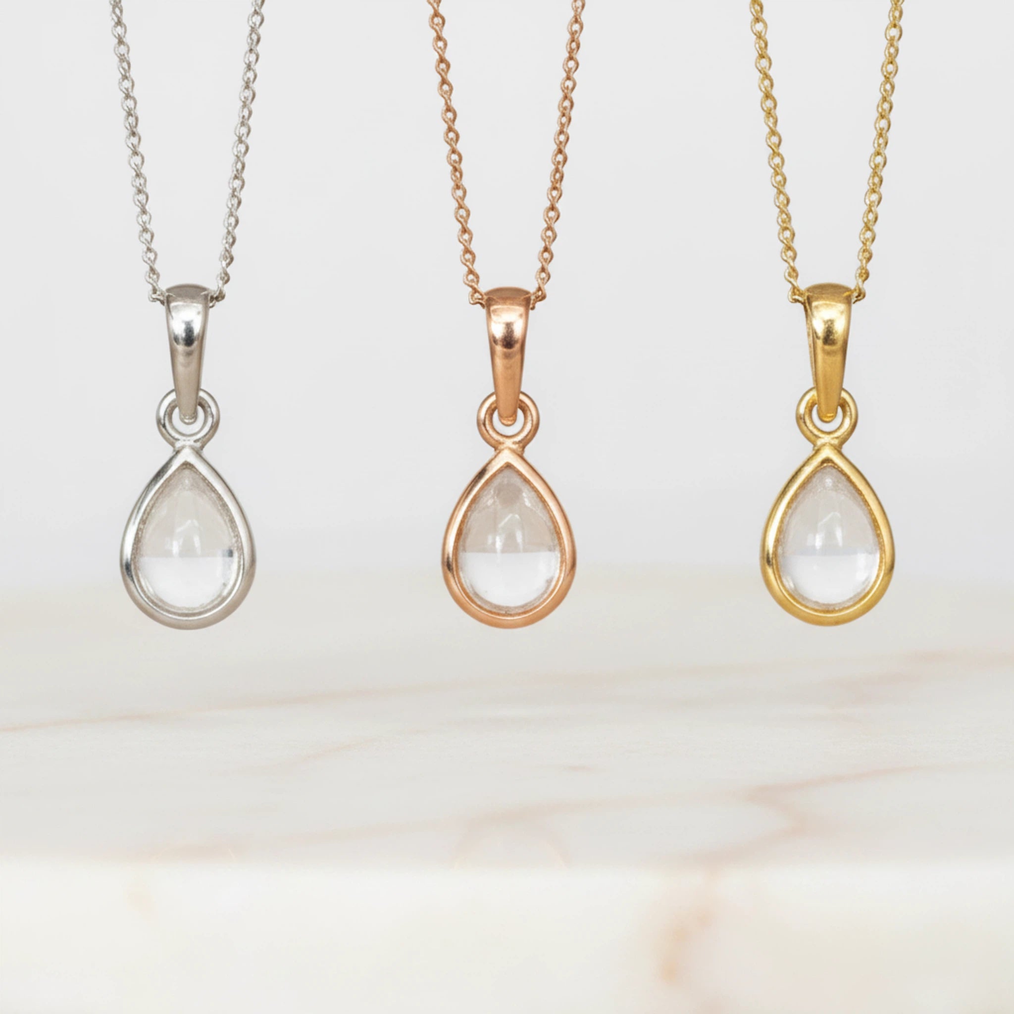Three April Birthstone Necklaces with teardrop-shaped natural Clear Quartz pendants in silver, rose gold and gold white background.