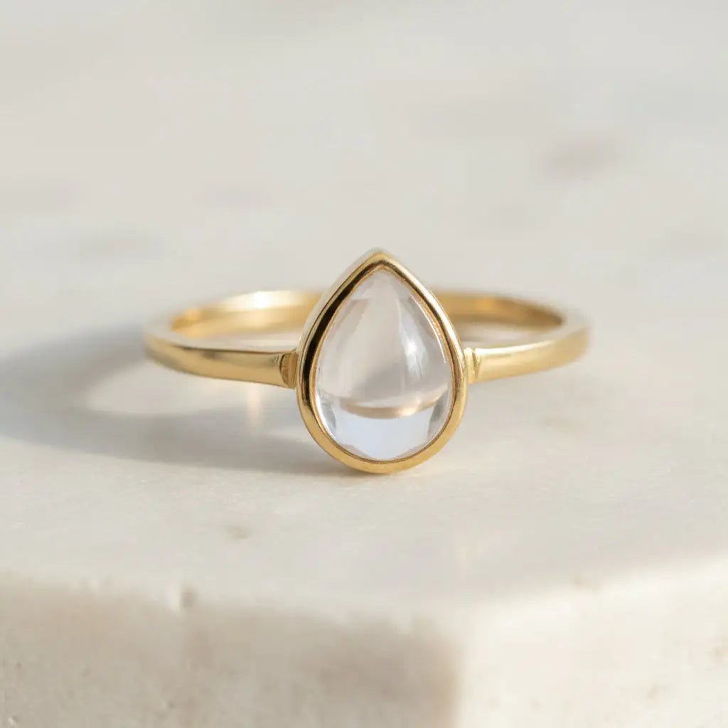 Front view of gold-plated April birthstone ring with pear-shaped clear quartz cabochon in a bezel setting on a light surface