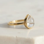 Gold April Birthstone Ring with a teardrop-shaped cabochon and bezel set natural clear quartz gemstone on a light background